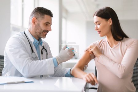 Photo for Male doctor holding syringe making vaccination injection dose in shoulder of young pregnant female patient. Flu vaccine clinical trials, virus treatment side effect - Royalty Free Image