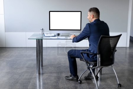 Téléchargez les photos : Vue arrière arrière de l'homme d'affaires en costume regardant l'ordinateur PC avec écran blanc pour modèle maquette, assis au bureau, espace de copie, pleine longueur - en image libre de droit