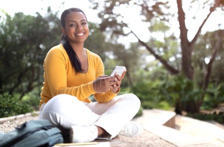 Téléchargez les photos : Joyeuse étudiante brésilienne utilisant son téléphone portable et envoyant des SMS en ligne, assise à l'extérieur dans un parc ou un campus universitaire, souriant à la caméra, copiant de l'espace. Étudiante excitée tenant smartphone - en image libre de droit