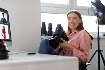 Téléchargez les photos : Jolie photographe positive travaillant dans le bureau du studio, assise au bureau avec ordinateur, tenant un appareil photo professionnel, vérifiant les photos, copiant l'espace - en image libre de droit