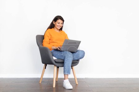 Photo for Cheerful beautiful young hindu lady wearing casual outfit sitting in armchair over white blank wall, surfing on Internet, chatting with friends, using modern pc laptop, copy space - Royalty Free Image