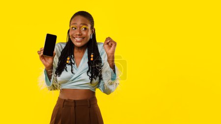 Foto de Oferta de comunicación móvil. Joyful African American Lady Mostrando Smartphone con pantalla vacía y haciendo gestos Sí en el fondo del estudio amarillo. Wow Aplicación Anuncio. Mockup, Panorama con espacio de copia - Imagen libre de derechos