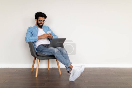 Photo for Online Communication. Smiling Indian Guy Using Laptop And Messaging On Smartphone While Resting In Chair Indoors, Happy Eastern Man Enjoying Modern Technologies And Digital Leisure, Copy Space - Royalty Free Image
