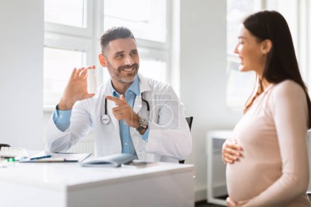 Foto de Prescripción de vitaminas. Médico varón sonriente demostrando recetar medicamentos a la mujer embarazada después del chequeo en la clínica. Amistoso terapeuta masculino apuntando a la botella con pastillas o vitaminas - Imagen libre de derechos