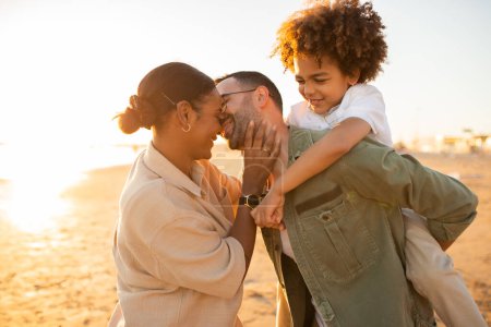 Photo for Young happy family spending time outdoors at the beach, spouses father and mother kissing, man piggybacking kid boy while walking by seaside - Royalty Free Image