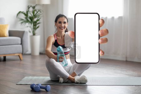 Photo for Nice fitness moble app concept. Cheerful pretty sporty young eastern woman sitting on yoga mat, holding bottle of water, showing smartphone with white empty screen, home interior, mockup - Royalty Free Image