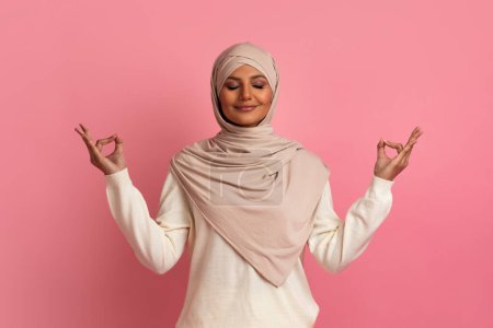Photo for Zen. Relaxed Peaceful Muslim Woman In Hijab Meditating With Closed Eyes, Calm Islamic Lady Coping With Stress, Practicing Yoga, Keeping Hands In Mudra Gesture While Posing On Pink Studio Background - Royalty Free Image