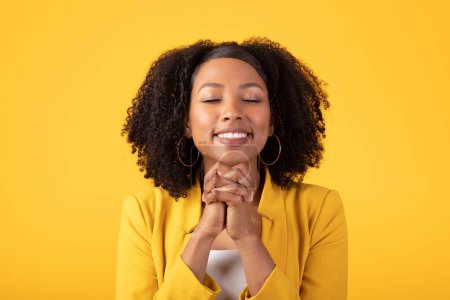 Téléchargez les photos : Joyeux sourire dame noire faire un geste de prière, faire un vœu, demander un miracle, debout isolé sur fond jaune studio. Espoir, rêves, émotions humaines - en image libre de droit