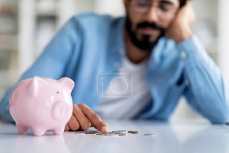 Photo for Lack Of Finances. Upset Indian Man Counting Coins Taken Out Of Piggy Bank, Depressed Eastern Male Sitting At Table At Home, Having Financial Crisis, Suffering Budget Planning Difficulties - Royalty Free Image