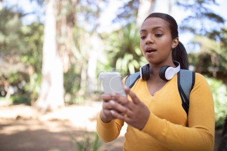 Photo for Shocked brazilian teen girl with backpack, reading message on smartphone with open mouth, standing outdoors in park. Reaction to news, app for study, knowledge, education - Royalty Free Image