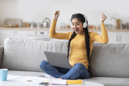 Photo for Great News. Happy Excited Indian Woman In Hedphones Celebrating Success With Laptop At Home, Overjoyed Young Eastern Lady Looking At Computer Screen, Raising Fists And Exclaining With Joy, Closeup - Royalty Free Image