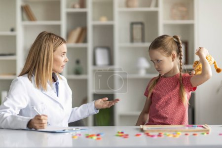 Photo for Aggressive Child Therapy. Cute Little Girl Feeling Angry During Therapy Session With Psychotherapist Lady, Upset Small Kid Threaten Specialist Doctor With A Toy, Having Temper Tantrum - Royalty Free Image