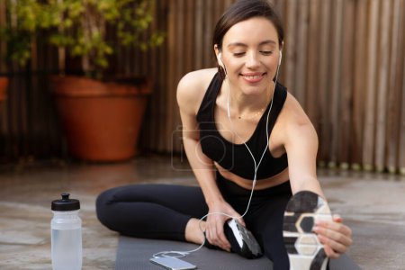 Photo for Glad young caucasian slender woman in sportswear and headphones doing stretching for legs on mat with bottle of water outdoor. Music for sports and warmup, workout, health care in city - Royalty Free Image