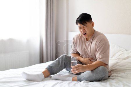 Téléchargez les photos : Malheureux malade chinois d'âge moyen assis sur le lit et touchant son ventre, souffrant de douleurs à l'estomac le matin, portant un pyjama, tenant un verre d'eau. Intoxication alimentaire, concept de diarrhée - en image libre de droit