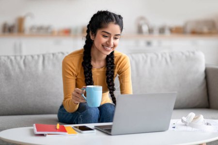 Foto de Estudio de la mujer india joven con el ordenador portátil y beber café en casa, sonriendo hembra oriental usando la computadora para la educación en línea y disfrutando de la bebida caliente mientras está sentado en el sofá en la sala de estar, espacio de copia - Imagen libre de derechos