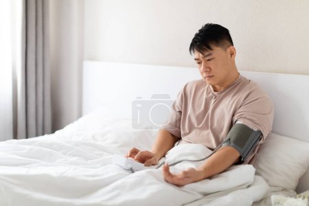 Foto de Hombre asiático de mediana edad preocupado vistiendo pijama sentado en la cama, comprobando su presión arterial con un moderno tonómetro electrónico. Hacer chequeo matutino después de despertarse o sentirse enfermo, copiar espacio - Imagen libre de derechos