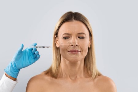 Photo for Woman looks at syringe, afraid of aesthetic surgery, grey studio background. Middle aged lady scared receiving hyaluronic acid injection on her face wrinkles fine lines, looking at needle - Royalty Free Image