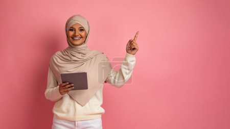 Photo for Online Offer. Happy Muslim Woman Holding Digital Tablet And Pointing At Copy Space With Finger, Smiling Arabic Woman In Headscarf Using Modern Gadget And Showing Free Place For Ad, Pink Background - Royalty Free Image