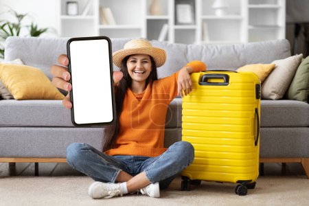 Photo for Cheerful pretty young arab lady wearing summer hat tourist sitting on floor by yellow luggage at home, showing smartphone with white blank screen, mockup, hot travelling offer, copy space - Royalty Free Image