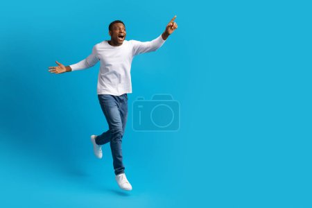 Foto de Bonita oferta. alegre guapo joven negro chico en casual hipster saltando y corriendo en el aire sobre azul estudio fondo, apuntando a espacio de copia para la publicidad, longitud completa - Imagen libre de derechos
