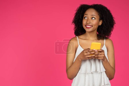 Foto de Encantada dama afroamericana milenaria reflexiva con teléfono mirando espacio libre aislado sobre fondo rosa, estudio. Enorme oferta y aplicación, buenas noticias, mensaje, venta y blog - Imagen libre de derechos