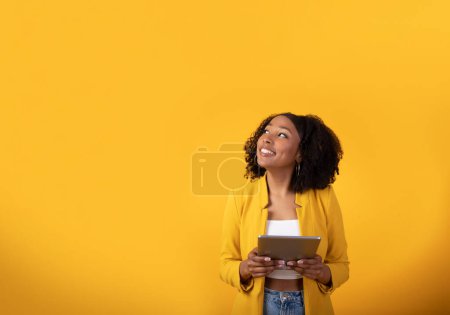 Téléchargez les photos : Heureuse jeune femme noire utilisant une tablette numérique sur fond de studio jaune, regardant l'espace de copie et souriant, vérifiant la plus récente application mobile, shopping, banque, étudier en ligne, bannière - en image libre de droit