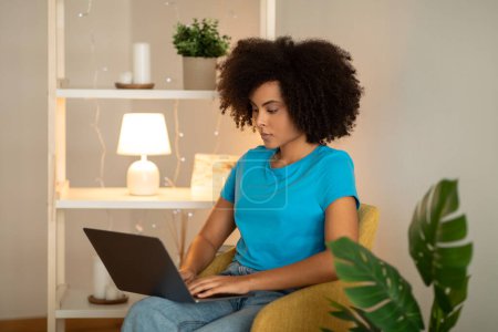 Photo for Concentrated millennial curly black lady student manager typing on computer in living room interior. Work, freelance, business and study remotely at home, device and social networks - Royalty Free Image