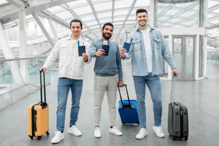 Geschäftsreisende Freunde. Glückliche junge Männer, die mit Reisegepäck, Pässen und Bordkarten im Abflugterminal des Flughafens posieren. Billige Tickets Anzeige. Volle Länge