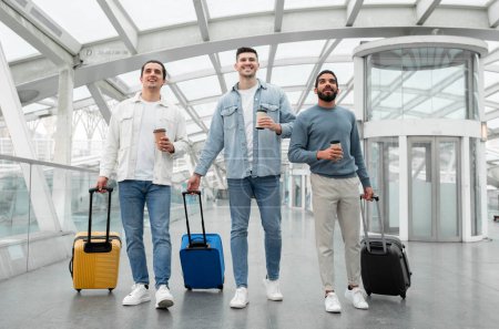 Téléchargez les photos : Globetrotting Vibe. Trois amis hommes marchant avec des valises de voyage tenant des tasses à café en papier, allant sur une nouvelle aventure de vacances ensemble posant dans l'aéroport moderne. Longueur totale - en image libre de droit