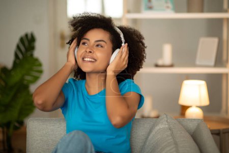 Photo for Portrait of satisfied millennial curly black woman in wireless headphones enjoy spare time, dreaming, has fun on sofa in cozy living room interior. Human listen music, audio app - Royalty Free Image