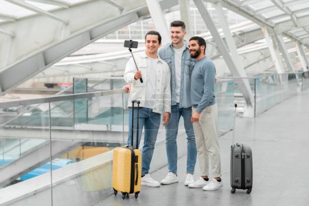 Photo for Pre-Boarding Pastime. Three Tourists Guys With Luggage Having Fun Making Selfie On Phone For Social Media Blog At Bus Station Or Airport Terminal Indoor. Friends Making Video For Travel Blog - Royalty Free Image