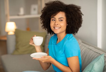 Foto de Encantada señora negra rizada milenaria sostener taza de té, disfrutar del tiempo libre, perezoso y comodidad en el sofá en el acogedor interior de la sala de estar. Pausa para tomar café por la noche, descansar, relajarse en casa el fin de semana - Imagen libre de derechos