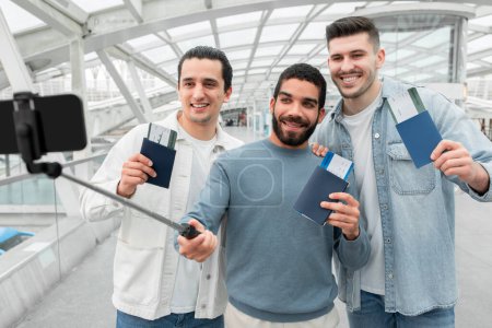 Photo for Travel Blog. Three Happy Tourists Men Making Selfie On Smartphone Enjoying Exciting Trip, Showing Passports And Boarding Passes Advertising Cheap Flight Offer In Modern Airport Deprature Terminal - Royalty Free Image