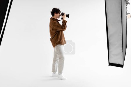 Téléchargez les photos : Vue latérale du jeune homme photographe prenant des photos à l'aide d'un appareil photo professionnel et d'un équipement d'éclairage dslr, debout sur fond blanc, espace de copie, pleine longueur - en image libre de droit