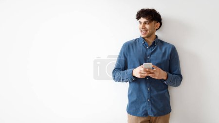 Photo for Mobile Offer. Cheerful Young Arab Guy Using Smartphone Texting And Looking Aside At Free Space Against White Studio Wall. Man Advertising Application For Cellphone. Panorama - Royalty Free Image