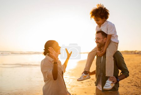 Photo for Sand, sea and smiles. Happy young diverse family of three spending free time outdoors, walking and having fun on seaside in the evening, father holding son on shoulders - Royalty Free Image