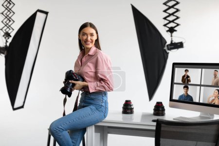 Photo for Happy creative photographer lady sitting on table workplace, holding her DSLR camera, smiling at camera while working with computer in modern photostudio - Royalty Free Image