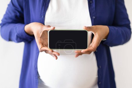 Photo for Millennial black woman in domestik clothes with big belly enjoys pregnancy, holds smartphone with empty screen. Health care, app for motherhood and sonography, blog and website, cropped - Royalty Free Image