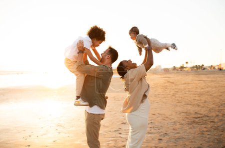 Photo for Coastal captures. Parents playing and having fun with their sons, throwing up boys in the air, spending time on the beach at sunset, side view, free space - Royalty Free Image