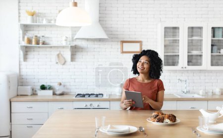 Téléchargez les photos : Smart Home Concept. Jeune femme noire contrôlant la lumière de la maison avec tablette numérique, femme afro-américaine utilisant un gadget moderne pour ajuster l'éclairage dans la cuisine, profitant d'un logiciel pour le confort domestique - en image libre de droit