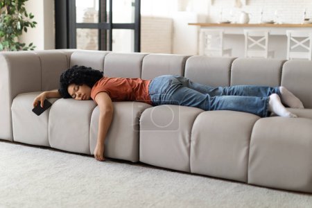 Téléchargez les photos : Concept d'épuisement. Jeune femme noire fatiguée dormant sur le canapé à la maison, Femme afro-américaine épuisée avec Smartphone dans la main enterré visage dans le canapé, sieste dans le salon, la chance de souffrir d'énergie - en image libre de droit