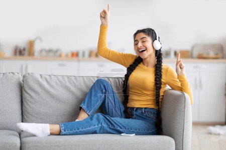 Photo for Domestic Fun. Cheerful Young Indian Woman Listening Music In Wireless Headphones While Relaxing On Couch At Home, Happy Eastern Female Dancing And Singing While Resting On Sofa In Living Room - Royalty Free Image