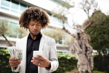 Photo for Serious black businessman using smartphone and holding dollar cash, having problems with online banking while standing outdoors in park near office building, free space - Royalty Free Image
