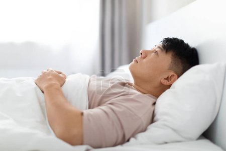 Photo for Frustrated handsome middle aged asian man lying in bed in the morning at home, staring ceiling, feeling trapped in current circumstances, lacking control, options for change, side view, copy space - Royalty Free Image