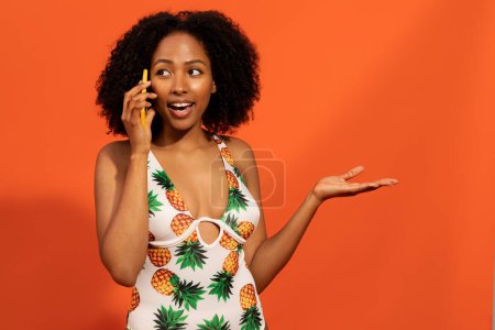 Téléchargez les photos : Joyeux émotionnel jeune femme afro-américaine avec les cheveux touffus en maillot de bain à la mode parler sur téléphone portable et gestuelle, regarder de côté, partager des nouvelles passionnantes sur fond de studio orange, copier l'espace - en image libre de droit