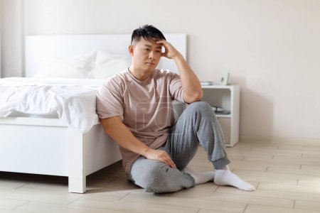 Photo for Sad middle aged asian man wearing pajamas sitting on bedroom floor at home, might have experienced significant loss, failed business, missed opportunity, personal setback, copy space - Royalty Free Image