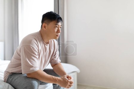 Foto de Pensativo hombre asiático de mediana edad con pijama sentado en la cama en casa, mirando el espacio de copia. Crisis existencial, cuestionamiento del sentido y propósito de la vida, sentirse insatisfecho o insatisfecho - Imagen libre de derechos