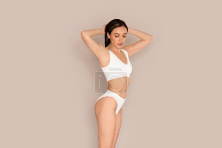 Photo for Body positivity. Slim well-fit pretty young brunette european woman wearing white comfy casual underwear demonstrating perfect body on beige studio background, holding arms behind head, copy space - Royalty Free Image