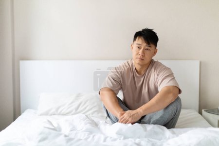 Photo for Upset handsome middle aged asian man sitting on bed at home, feeling down, looking at copy space, struggling with depression, anxiety, or other mental health conditions - Royalty Free Image