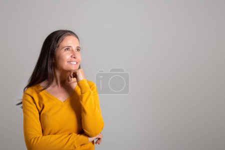 Foto de Sonriendo pensativa mujer europea bastante vieja mira el espacio vacío, piensa en el anuncio y la oferta, disfrutar de las compras en fondo gris estudio. Elija, venta enorme, crear idea y estilo de vida - Imagen libre de derechos